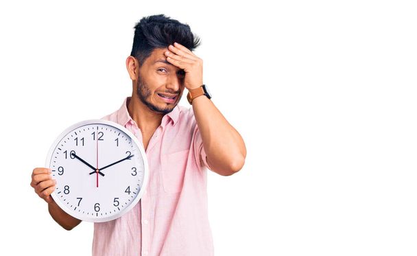 Handsome latin american young man holding big clock stressed and frustrated with hand on head, surprised and angry face  - Foto, Imagem