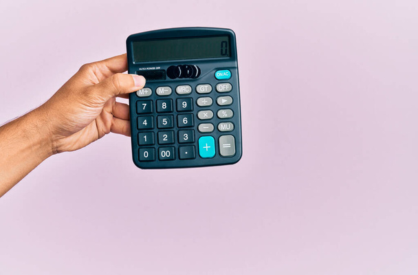 Mão de jovem hispânico homem segurando calculadora sobre fundo rosa isolado. - Foto, Imagem