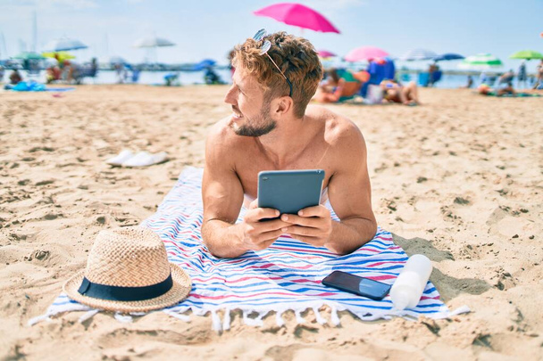 Bellissimo uomo caucasico fitness in spiaggia in una giornata di sole sdraiato sull'asciugamano utilizzando il dispositivo touchpad - Foto, immagini