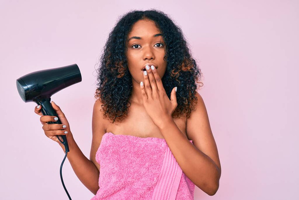 Mladá afro-americká žena ve sprchovém ručníku se sušičkou zakrývající ústa rukou, šokovaná a vystrašená z omylu. překvapený výraz  - Fotografie, Obrázek