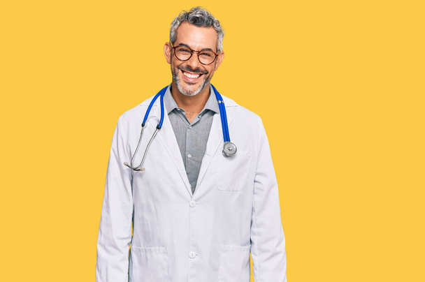 Middle age grey-haired man wearing doctor uniform and stethoscope looking positive and happy standing and smiling with a confident smile showing teeth  - Photo, Image