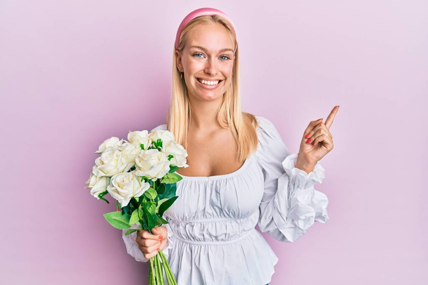 Nuori blondi tyttö tilalla kukkia hymyilevä onnellinen osoittaa käsi ja sormi sivulle  - Valokuva, kuva