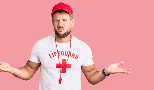 Young caucasian man wearing lifeguard t shirt holding whistle clueless and confused with open arms, no idea concept.  - Photo, Image