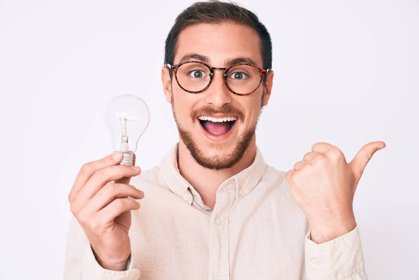 Young handsome man holding lightbulb for inspiration and idea pointing thumb up to the side smiling happy with open mouth  - Foto, Bild