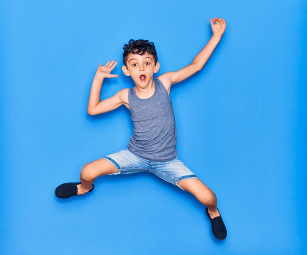 Αξιολάτρευτο παιδί που φοράει καθημερινά ρούχα πηδώντας πάνω από απομονωμένο μπλε φόντο - Φωτογραφία, εικόνα