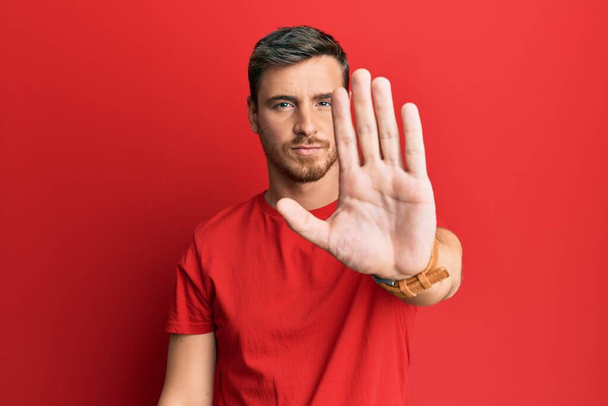 Красивый белый мужчина в обычной красной футболке делает остановку петь с ладонью руки. предупреждающее выражение с негативным и серьезным жестом на лице.  - Фото, изображение