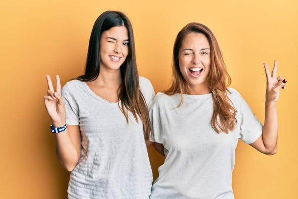 Famille hispanique de mère et fille portant un t-shirt blanc décontracté souriant avec le visage heureux clignant à la caméra faisant signe de victoire avec les doigts. numéro deux.  - Photo, image
