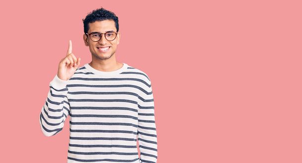 Hispanic handsome young man wearing casual clothes and glasses showing and pointing up with finger number one while smiling confident and happy.  - Photo, Image