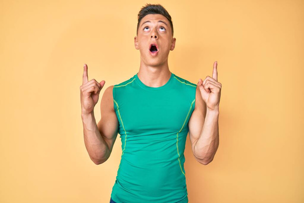 Mladý hispánec ve sportovním stylu s košilí bez rukávů užaslý a překvapený vzhlíží a ukazuje prsty a zvednutými pažemi.  - Fotografie, Obrázek