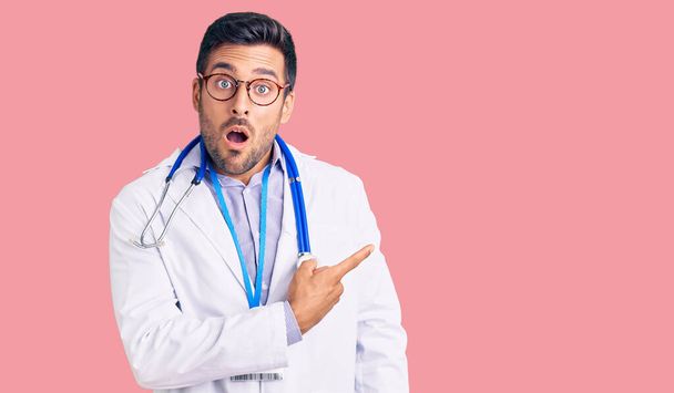Joven hombre hispano vistiendo uniforme médico y estetoscopio sorprendido señalando con el dedo hacia un lado, boca abierta expresión asombrada.  - Foto, imagen