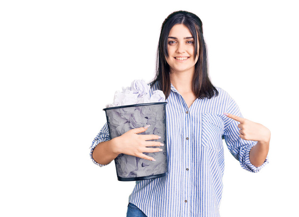Молодая красивая девушка держит в руках бумажную корзину, полную скомканных бумаг, указывая пальцем на себя улыбающейся счастливой и гордой  - Фото, изображение