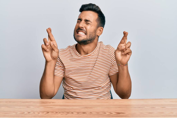 Een knappe Spaanse man in casual kleren op de tafel met een gekruiste vinger die glimlacht met hoop en ogen dicht. geluk en bijgelovig concept.  - Foto, afbeelding