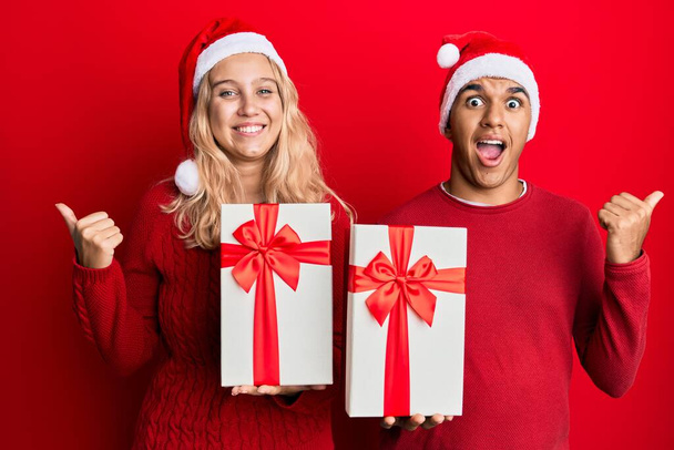 Młoda para międzyrasowa w świątecznym kapeluszu i trzymająca prezent z kciukiem w górze, uśmiechnięta szczęśliwa z otwartymi ustami  - Zdjęcie, obraz