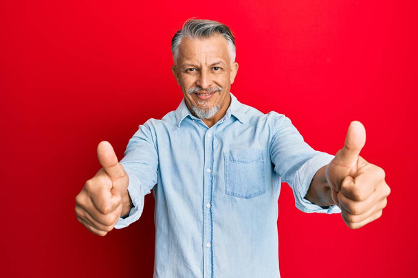 Чоловік середнього віку у сіро-волосистому одязі, що схвалює позитивний жест рукою, великі пальці посміхаються і щасливі за успіх. жест переможця
.  - Фото, зображення