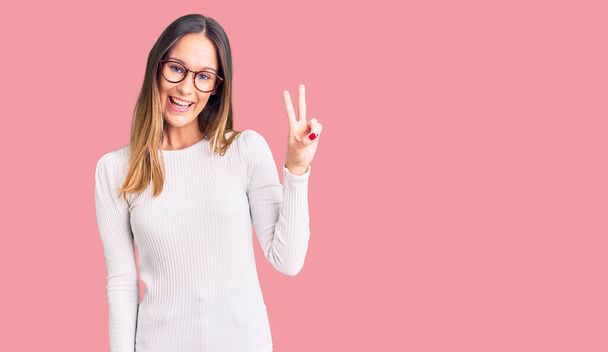 Beautiful brunette young woman wearing casual white sweater and glasses smiling with happy face winking at the camera doing victory sign with fingers. number two.  - Фото, зображення