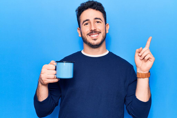 Young hispanic man holding coffee smiling happy pointing with hand and finger to the side  - Valokuva, kuva