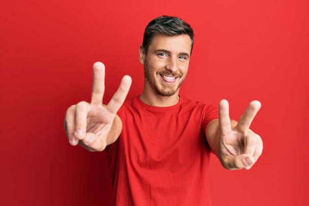 Knappe blanke man met een casual rood tshirt die glimlacht met zijn tong en vingers laat zien van beide handen die een overwinningsteken doen. nummer twee.  - Foto, afbeelding