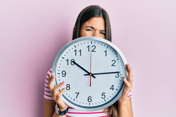 Jeune femme brune tenant une grande horloge couvrant le visage expression désemparée et confuse. concept de doute.  - Photo, image