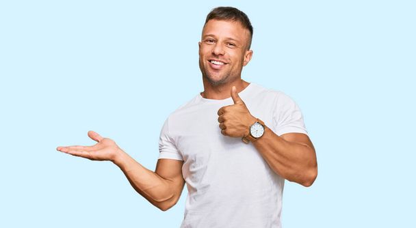Hezký svalovec v neformální bílé tričko ukazující dlaň a dělá v pořádku gesto s palci nahoru, usmívá šťastný a veselý  - Fotografie, Obrázek