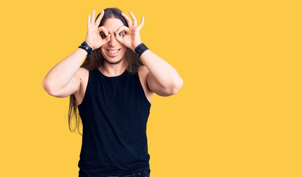 Young adult man with long hair wearing goth style with black clothes doing ok gesture like binoculars sticking tongue out, eyes looking through fingers. crazy expression.  - Fotografie, Obrázek