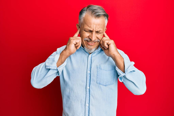 Middle age grey-haired man wearing casual clothes covering ears with fingers with annoyed expression for the noise of loud music. deaf concept.  - 写真・画像