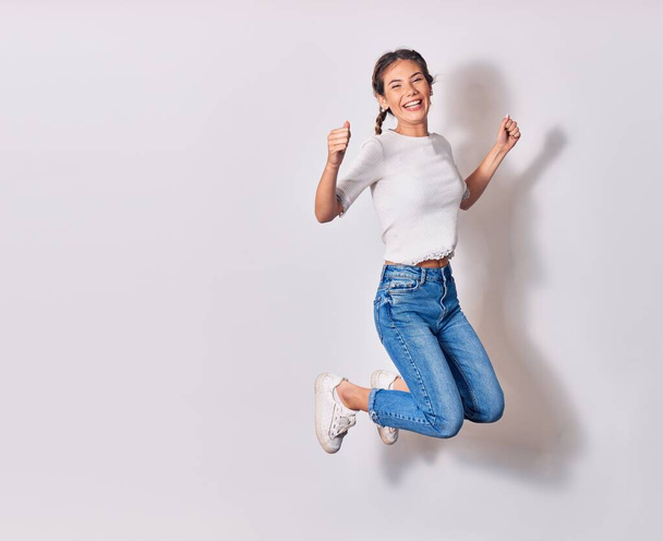 Young beautiful woman with braids wearing casual clothes smiling happy. Jumping with smile on face doing winner sign with fists up over isolated white background - Photo, Image