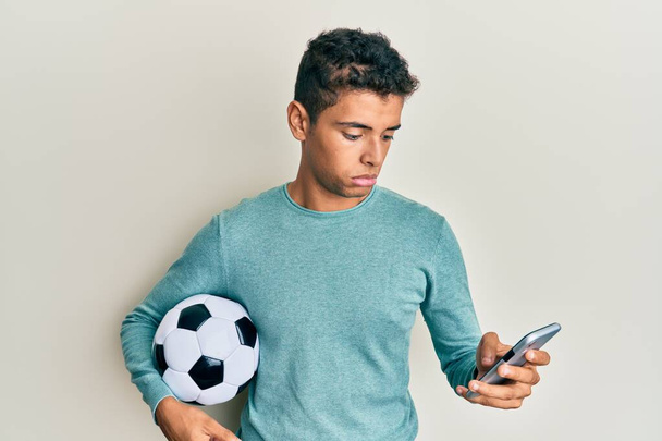 サッカーボールを持っている若いハンサムなアフリカ系アメリカ人男性は、スマートフォンを見て落ち込んでおり、苦痛のために心配し、怒りと恐怖を泣いています。悲しい表情.  - 写真・画像