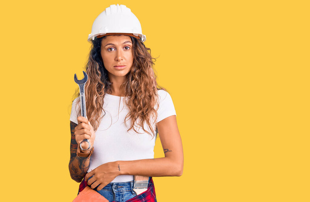 Young hispanic woman with tattoo wearing hardhat and builder clothes holding wrench thinking attitude and sober expression looking self confident  - Photo, Image