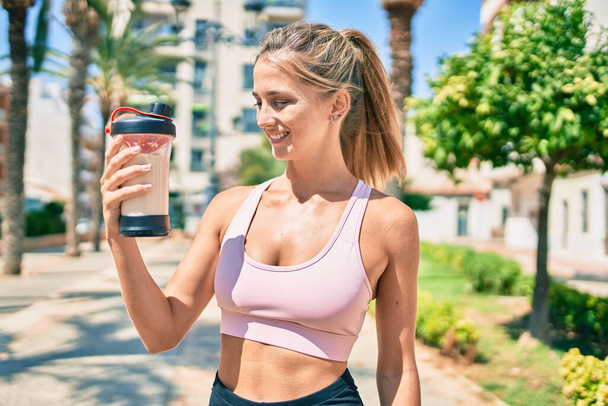 Young blonde sporty girl smiling happy drinking healthy protein smoothie at street of city. - Photo, Image