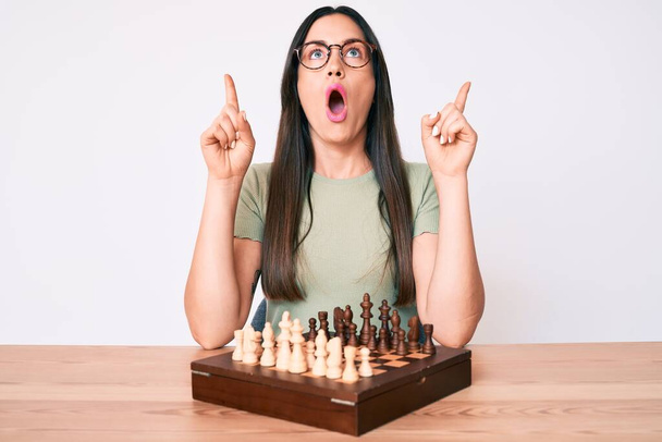 Young caucasian woman sitting at the desk playing chess amazed and surprised looking up and pointing with fingers and raised arms.  - Foto, Bild