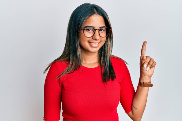 Young latin girl wearing casual clothes and glasses smiling with an idea or question pointing finger up with happy face, number one  - Photo, Image