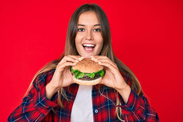Schöne kaukasische Frau isst einen leckeren klassischen Burger lächelnd mit einem fröhlichen und kühlen Lächeln auf dem Gesicht. Zähne zeigen.  - Foto, Bild