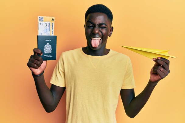 Giovane uomo afroamericano in possesso di aereo di carta e passaporto attaccare lingua fuori felice con espressione divertente.  - Foto, immagini