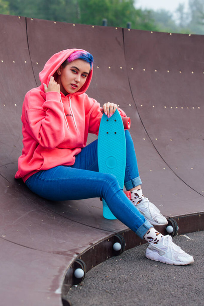 Stylish young woman with short colored hair sitting with her blue plastic skateboard in skatepark. Youth concept. - Fotografie, Obrázek