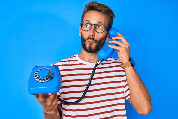 Handsome caucasian man with beard holding vintage telephone making fish face with mouth and squinting eyes, crazy and comical.  - Photo, Image
