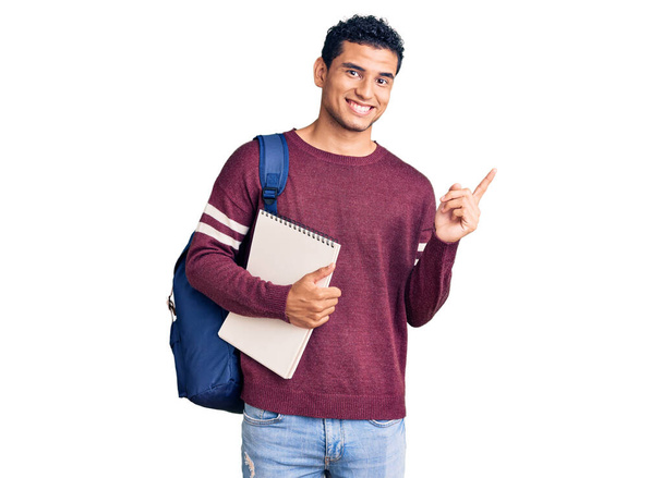 Hispanique beau jeune homme portant un sac à dos étudiant et un cahier souriant heureux pointant avec la main et le doigt sur le côté  - Photo, image