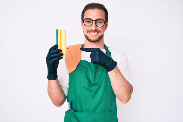 Young handsome man wearing apron holding scourer smiling happy pointing with hand and finger  - Photo, Image