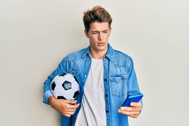 Hombre caucásico guapo sosteniendo pelota de fútbol mirando teléfono inteligente relajado con expresión seria en la cara. simple y natural mirando a la cámara.  - Foto, Imagen
