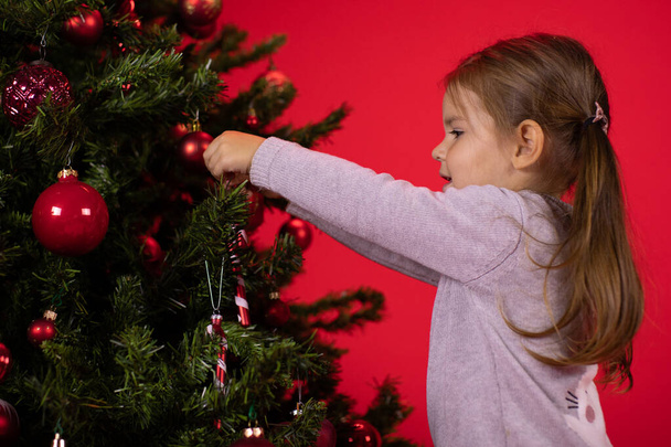baby girl hanging decorative toy on Christmas tree, red studio Xmas background - Foto, Imagen