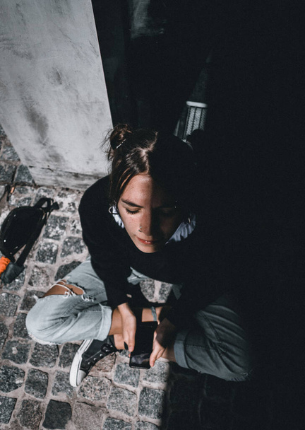 Ένα κάθετο πλάνο υψηλής γωνίας μιας νεαρής στιλάτης γυναίκας που κάθεται πάνω σε πλακόστρωτη πέτρα με μια σκιά που καλύπτει το μισό της πρόσωπο. - Φωτογραφία, εικόνα