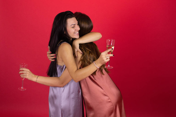 Две юные лесбиянки в элегантных платьях обнимаются, держа шампанское, красный фон - Фото, изображение