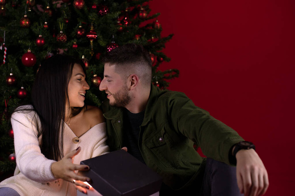 Молодая любящая пара, открывая долгожданный новогодний подарок, сидела рядом с елкой - Фото, изображение