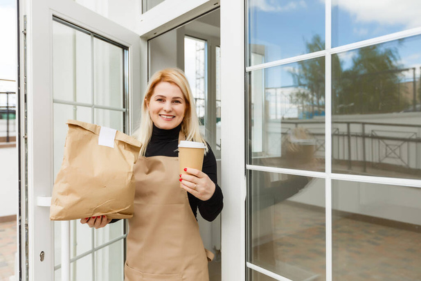 Mooie jonge vrouw in schort en leunend tegen de deuropening van muur koffiehuis of restaurant als eigenaar of werknemer - Foto, afbeelding