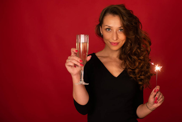 Young successful woman with champagne and sparkler on red background, copy space - Photo, Image