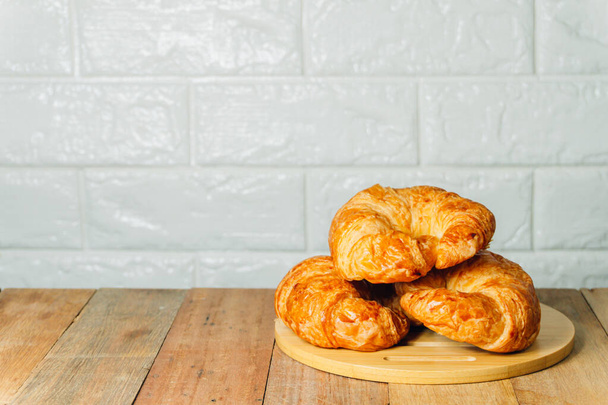 Drei hausgemachte Croissants stehen auf einem Holztablett, das auf einem Holztisch vor weißem Backsteinhintergrund steht.. - Foto, Bild