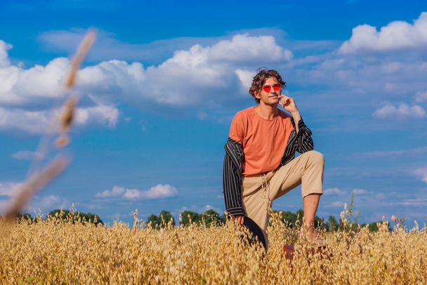 Rural Countryside Scene. Tall handsome man dressed in a black shirt, black hat and pink sunglasses standing with one leg on a brown vintage leather suitcase at golden oat field. Summer landscape with blue sky - Фото, зображення