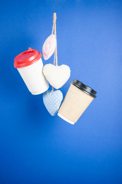 wegwerppapier vliegende koffiebekers met textiel harten op een blauwe achtergrond opknoping op. Afhaalkoffie concept. Abstracte achtergrond - Foto, afbeelding