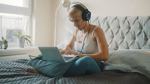Giovane donna bionda caucasica con auricolare gioco sul computer portatile nella sua camera da letto - Foto, immagini