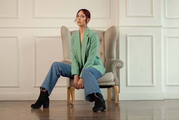 Portrait of a young girl with red hair gathered in a bunch with makeup and in a green jacket and jeans sits on an armchair in an empty room - Photo, Image