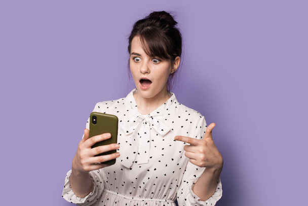 Brunette woman in a dress is pointing amazed at her phone screen posing with opened mouth on a violet studio wall - 写真・画像